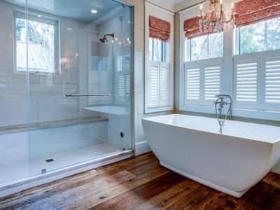 Why Now is the best time for bathroom renovations in Madisonville LA with Northshore Renovations and Contracting image of large walk in shower with glass doors next to large tub in front of windows with white wooden shutters and red curtains
