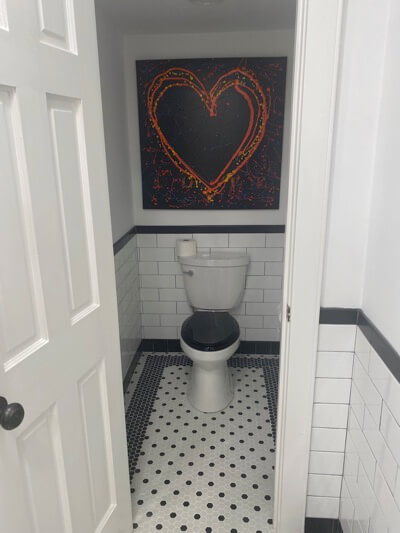 toilet in closet space of bathroom for privacy with black and white tile walls and floor with abstract heart painting hung above
